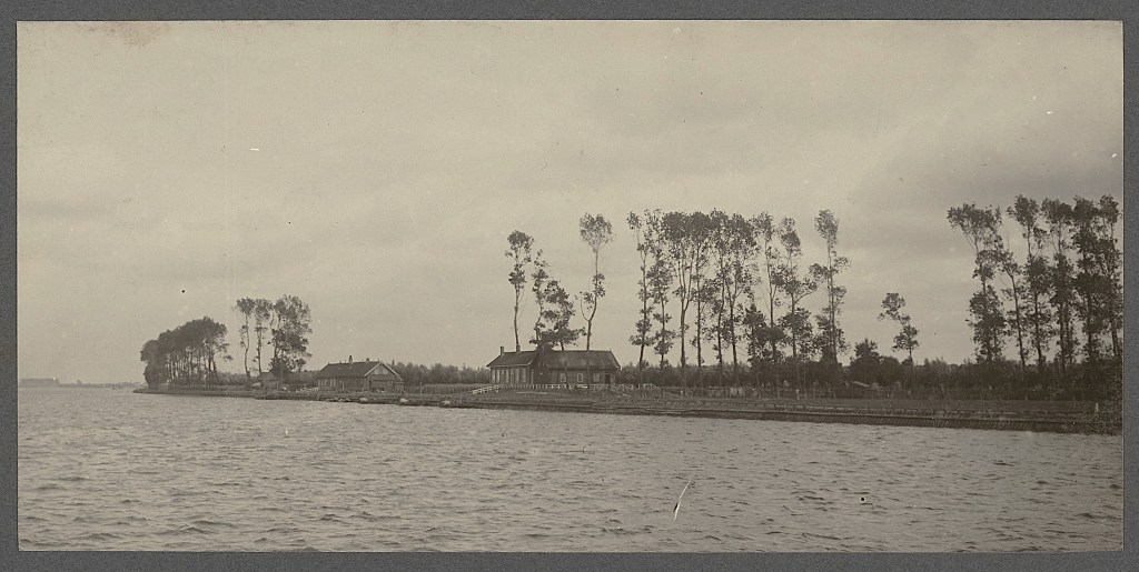 Oude zwart-wit foto van zalmvisserij Oranje Nassau, genomen vanaf het water. Een aantal gebouwtjes zijn zichtbaar en een rij populieren.